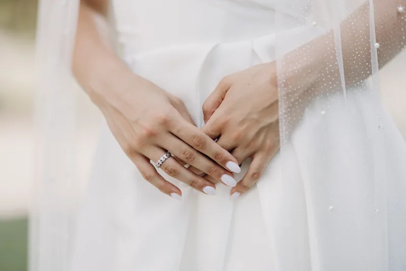 Close up of a bride's hands resting over her white dress, showing her wedding ring and soft veil.