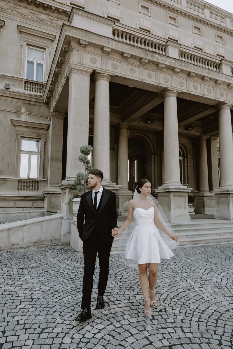 A couple hold hands walking across cobblestones before a grand columned building, the bride in a short dress.