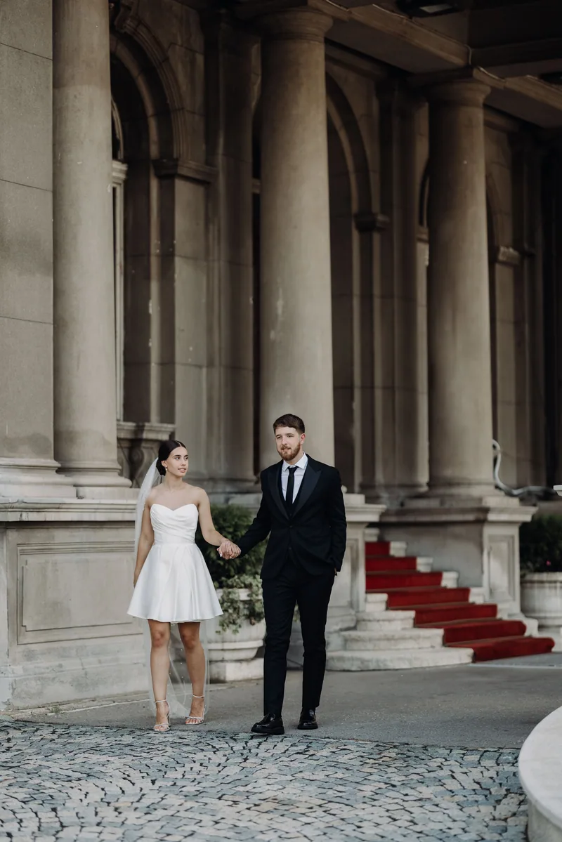 A couple walk hand in hand beneath an arched colonnade beside a red carpeted staircase.