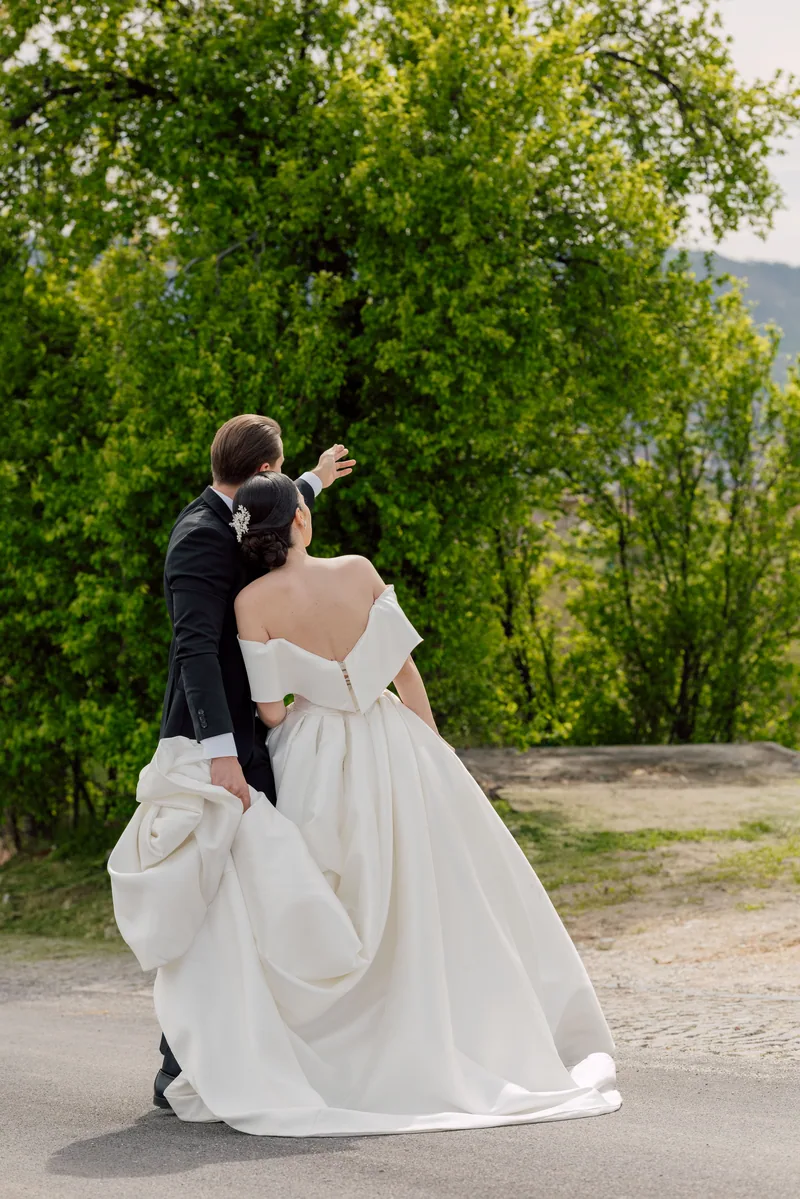 Seen from behind, a groom holds up the bride's long train as they walk toward lush green trees.