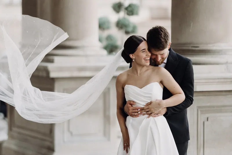 A bride laughs as her groom embraces her from behind and her long pearl veil floats out in the wind.